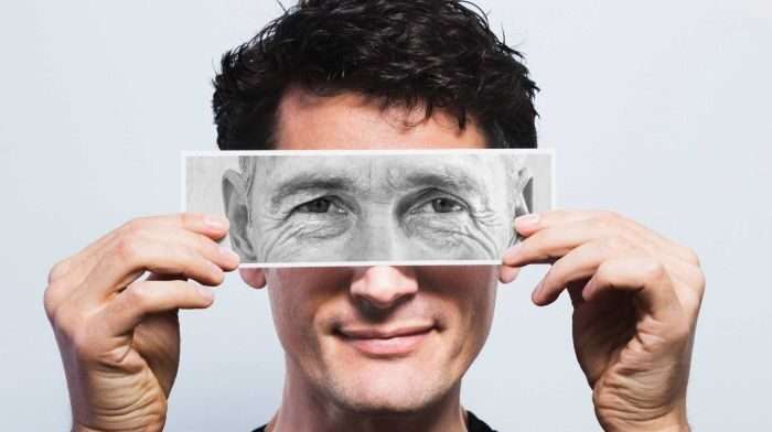 A young person holding a photograph in front of his eyes which shows the wrinkled eyes of an elderly man.